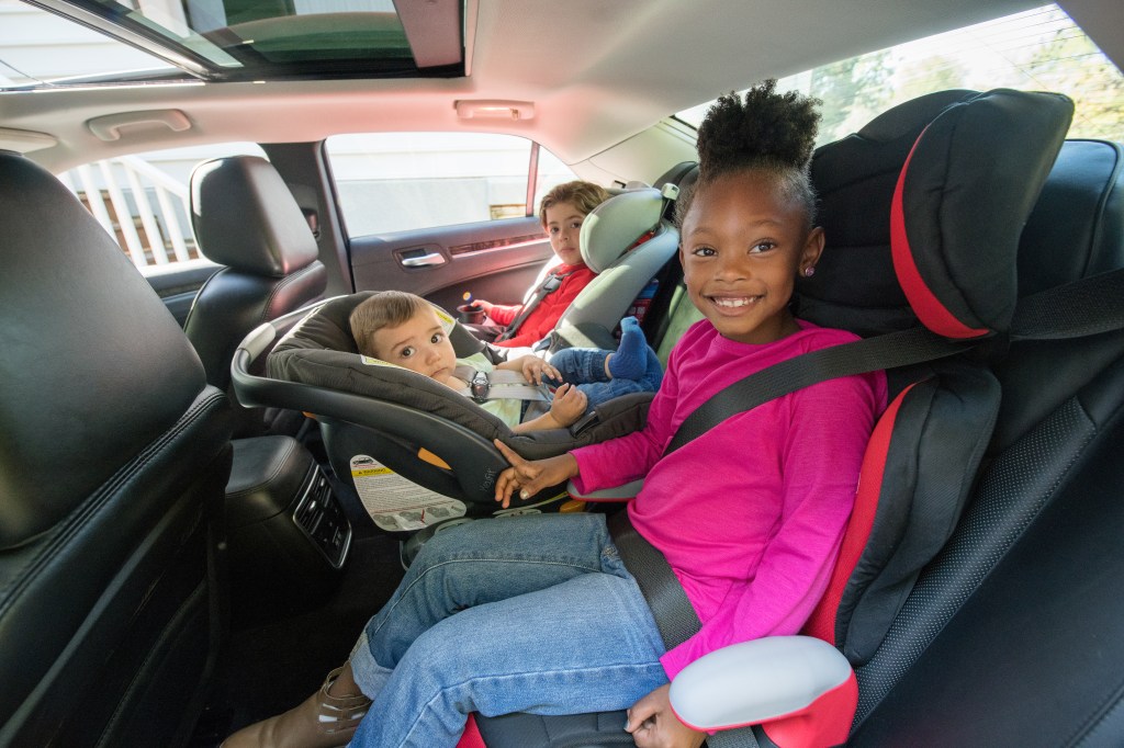 Photo of three children in car seats, sitting in the back seat of a vehicle. A young black girl is in a booster seat, a white infant is in a rear-facing car seat, and a young white boy is in a forward-facing car seat. 