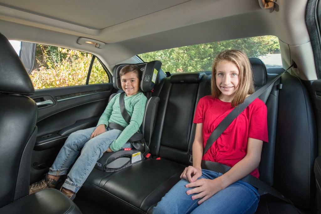 Two white children in the back seat of a vehicle. A young boy in a booster seat and a tween girl in a seat belt. 
